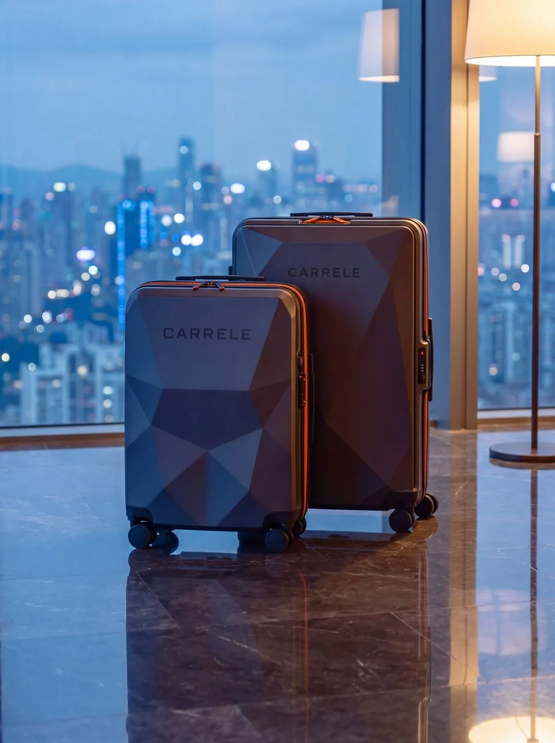 Two faceted graphite Carrele suitcases with orange zips by a window overlooking a city at dusk.