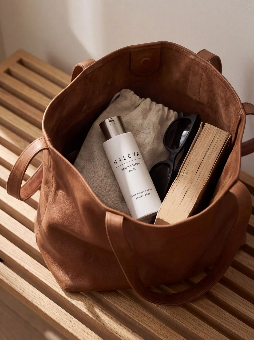 Halcya Barrier Serum in an open tan leather tote with sunglasses and a paperback, on a sunlit slatted wooden bench.